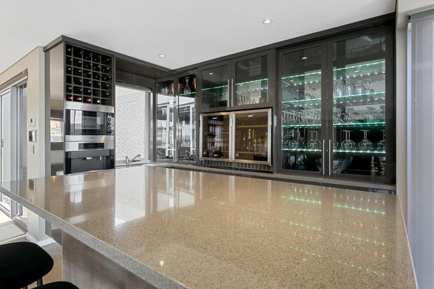 A contemporary kitchen and entertaining area dominated by a large polished stone island benchtop. Dark built-in cabinetry includes a wine rack, integrated appliances, illuminated glass-front display cabinets, and a compact beverage or bar station. The room has light walls, recessed ceiling lighting, extensive glazing, and a refined high-contrast palette of dark cabinetry, pale stone, and stainless-steel appliances.
