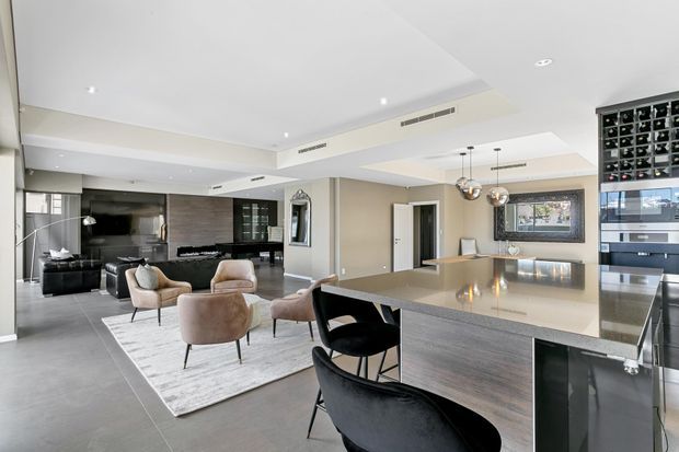 Open-plan contemporary kitchen and living area with a large stone island and integrated seating in the foreground, dark fixed cabinetry and a built-in wine display at right, and a furnished lounge area beyond. The space has light neutral finishes, recessed lighting, expansive glazing, and a refined monochromatic palette.