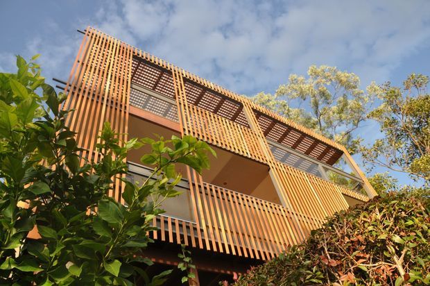 Low-angle exterior view of a multi-storey residential building largely screened by warm timber vertical battens. Open balcony and upper-level areas feature patterned timber lattice and dark-framed glazing, with substantial surrounding vegetation and a partly cloudy sky.