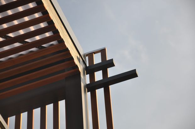Upward-looking architectural detail of a contemporary building exterior, featuring strong dark timber or steel horizontal beams and vertical supports forming a pergola-like overhang. Warm sunlight highlights the structure against a pale sky, creating a graphic, minimalist composition.