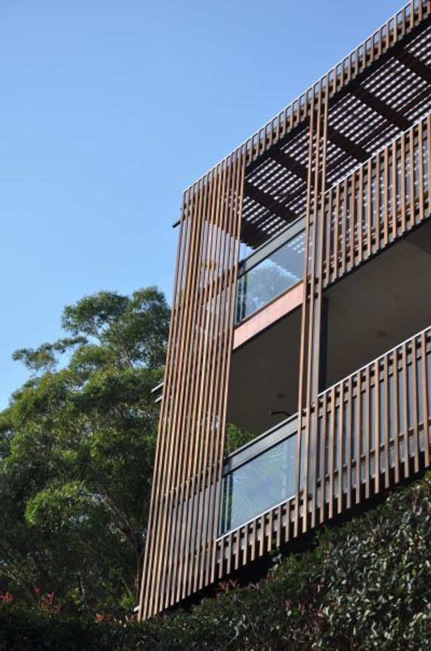 Upward-looking exterior view of a contemporary multi-level building integrated into a leafy setting. The facade is defined by extensive vertical timber-look screening, projecting balconies with clear glass balustrades, dark structural elements, and a slatted pergola or roof canopy above. Bright blue sky and mature vegetation surround the building.
