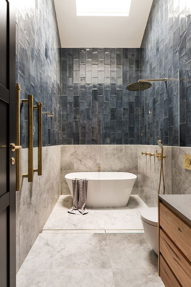 A narrow, refined bathroom with a freestanding white bathtub set on a raised tiled platform. The walls combine large pale stone-look tiles with vertically arranged dark blue-grey textured tiles, while brass shower fittings and hardware add warmth. A skylight provides natural light, and a timber vanity is partially visible at the right.