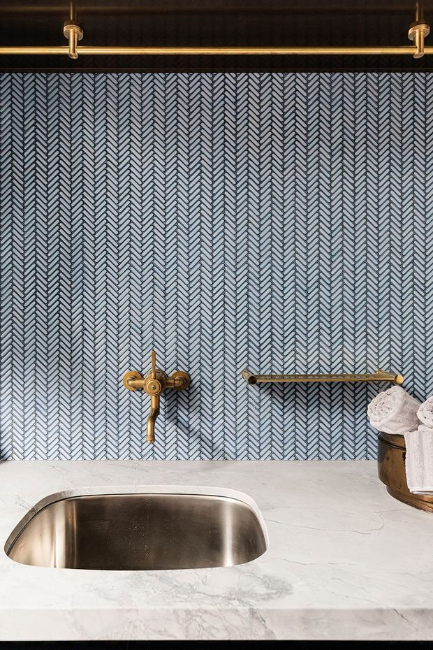 Close-up of a refined bathroom vanity featuring a white marble-look countertop with an integrated rounded rectangular basin, a blue-grey herringbone patterned tile splashback, and brushed brass wall-mounted tapware, shelf rail, and overhead hanging rail. Towels and a small container sit at the right edge.
