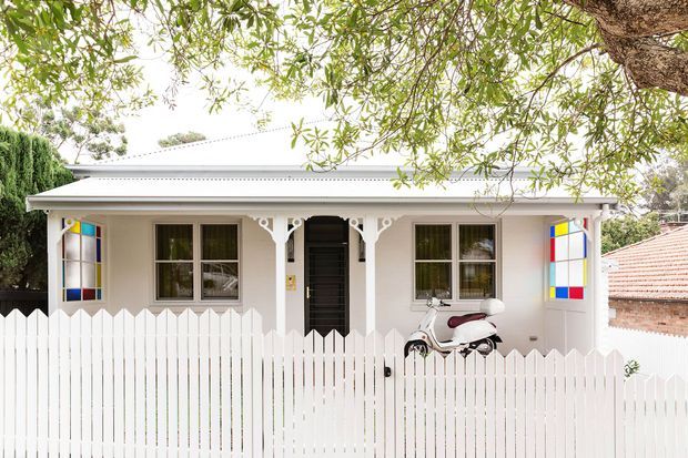 Street-facing view of a compact single-storey cottage with a pale rendered facade, low-pitched light metal roof, decorative white verandah posts and fretwork, colourful stained-glass window panels, and a white picket fence enclosing the front garden. A scooter is parked behind the fence, with mature trees framing the property.