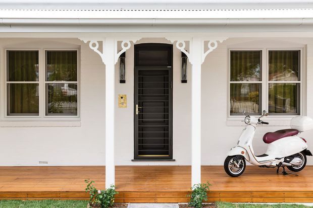 Front facade of a small traditional-style home with a white-painted exterior, decorative fretwork brackets, covered veranda, timber decking, black framed entry door, and symmetrical windows. A white scooter is parked on the veranda.