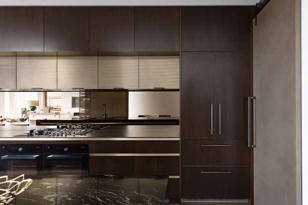 A sophisticated modern kitchen featuring extensive dark timber cabinetry, integrated tall storage and refrigerator units, a large central island with a dramatic dark veined stone surface, gas cooktop, built-in ovens, and a reflective backsplash. Horizontal window blinds and warm, restrained lighting create a refined, moody atmosphere.