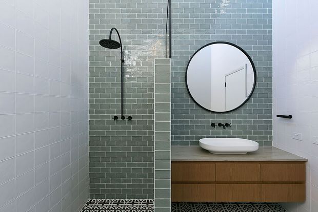 Contemporary bathroom featuring a walk-in shower with muted green subway tiles, a round black-framed mirror, floating timber vanity with a white vessel basin, matte black tapware, and patterned floor tiles.