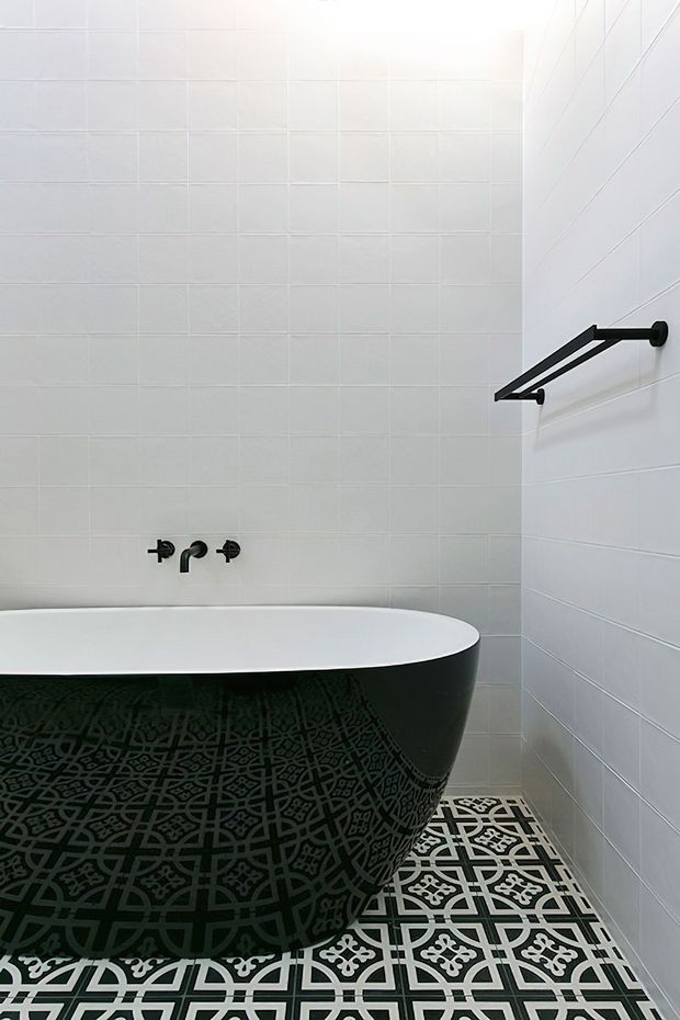 A compact, bright bathroom featuring a freestanding black bathtub with an ornate geometric patterned exterior, white tiled walls, patterned black-and-white floor tiles, and matte black wall-mounted tapware and towel rail.
