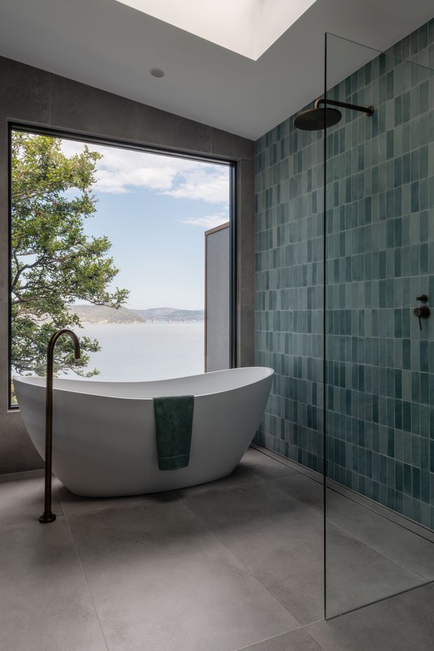 A refined contemporary bathroom with a freestanding white bathtub beside a large window overlooking a calm waterfront landscape. The room features large grey floor tiles, dark grey wall finishes, a teal-green vertical tile feature wall, black-bronze shower and bath fittings, a frameless glass shower screen, and a skylight that brings in natural light.