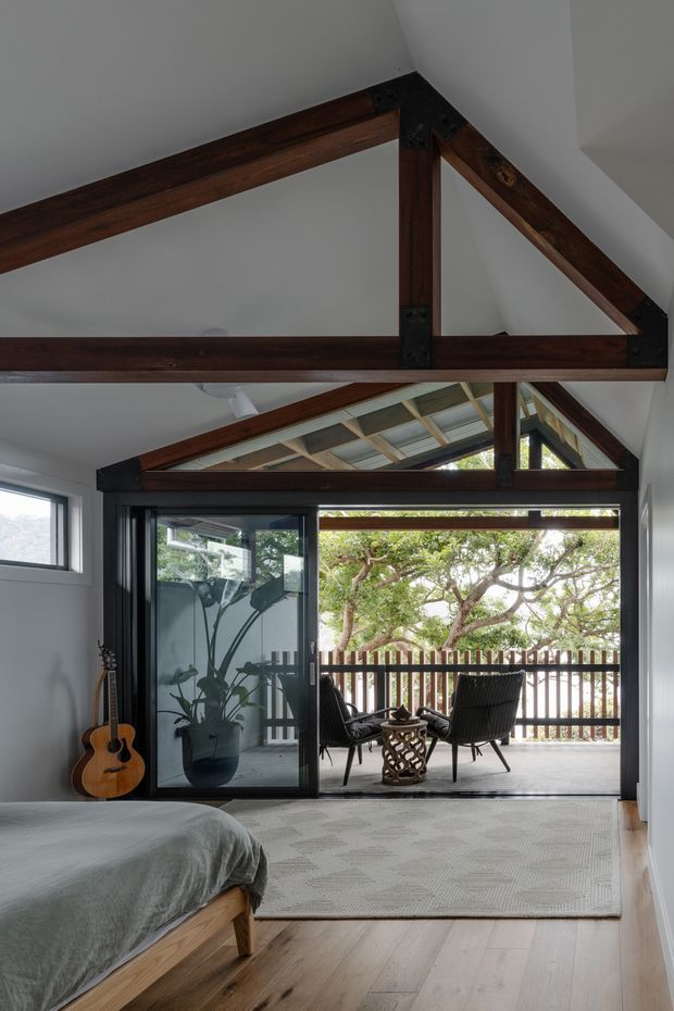 A bright bedroom opens directly onto a covered timber-framed outdoor terrace through large glazed sliding doors. The room features exposed dark timber structural beams and triangular roof trusses, white walls, pale timber flooring, a low timber bed with grey bedding, and a neutral patterned rug. A guitar and potted plant sit near the glazing, while outdoor lounge chairs and a timber balustrade are visible on the terrace beyond.