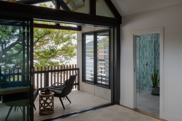 Interior view of a bright modern living area opening onto a covered balcony or terrace through large black-framed sliding glass doors. The space features exposed dark timber beams, pale walls, a light floor, and a lounge chair with a small side table. A bathroom is visible through the doorway, with blue-green tiled walls and a potted plant. The glazing and balcony overlook leafy surroundings.