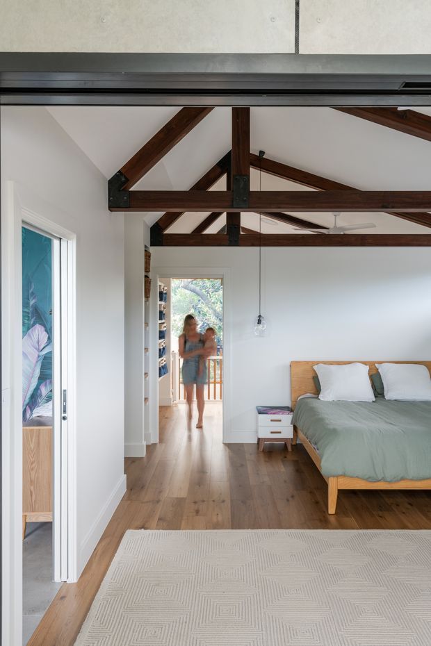 Bright contemporary bedroom with a timber-framed pitched ceiling and exposed dark wood beams, white walls, warm timber flooring, and a simple timber bed dressed in pale green and white bedding. A large textured neutral rug, pendant lights, and a small bedside table complete the room. An open doorway leads to a hallway and balcony, while a partial adjoining room is visible at left.