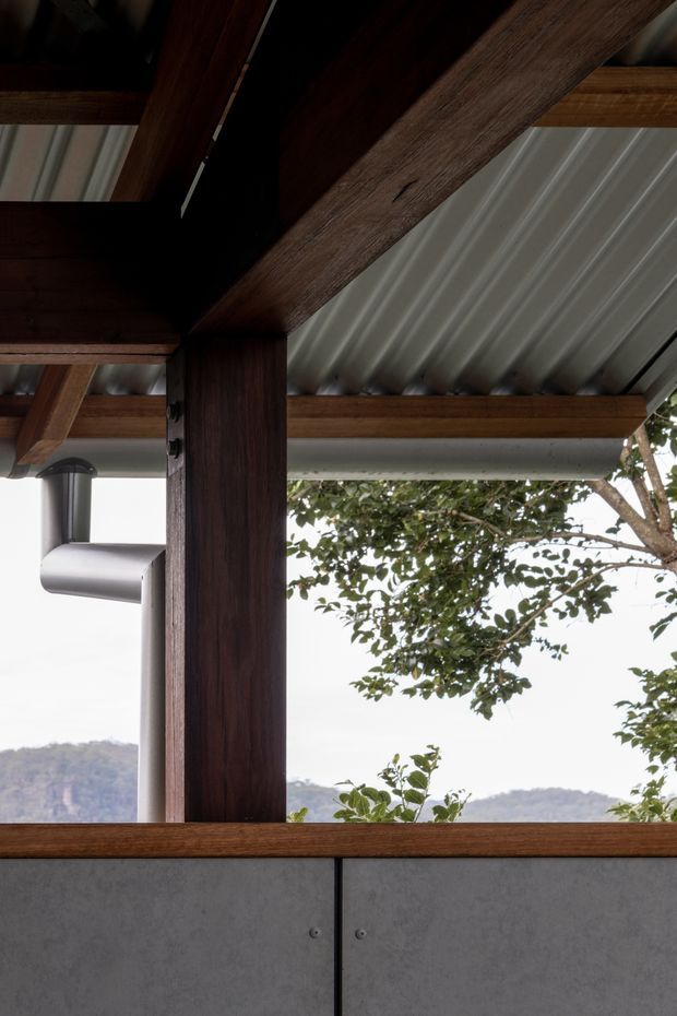 Close-up architectural detail of a covered outdoor structure, showing dark timber posts and exposed beams supporting a corrugated metal roof, with a grey panelled lower wall, visible roof drainage, and a leafy landscape beyond. The composition emphasises robust post-and-beam construction and natural timber against grey metal and concrete-like surfaces.