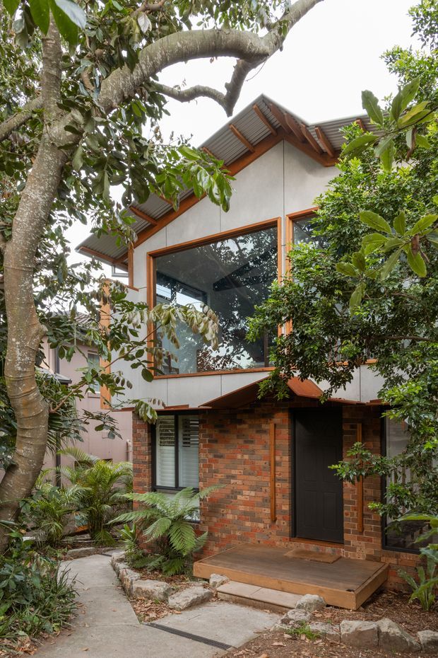 A two-storey residential building nestled in dense mature greenery. The facade combines red and brown brickwork at the lower level with light rendered or panelled upper walls, warm timber-framed glazing, and a pitched gabled roof with exposed timber eaves and corrugated metal roofing. A small covered entry porch opens onto a timber landing, with a narrow concrete garden path and lush fern-filled landscaping leading to the entrance.