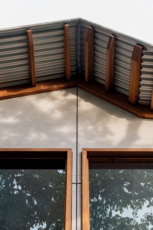 Close-up exterior architectural detail showing a corrugated metal wall and roof-edge junction with exposed dark timber framing and trim. Two large timber-framed glazed openings occupy the lower portion, reflecting surrounding trees. The composition highlights the contrast between grey ribbed metal cladding, warm timber, and concrete-coloured wall panels.