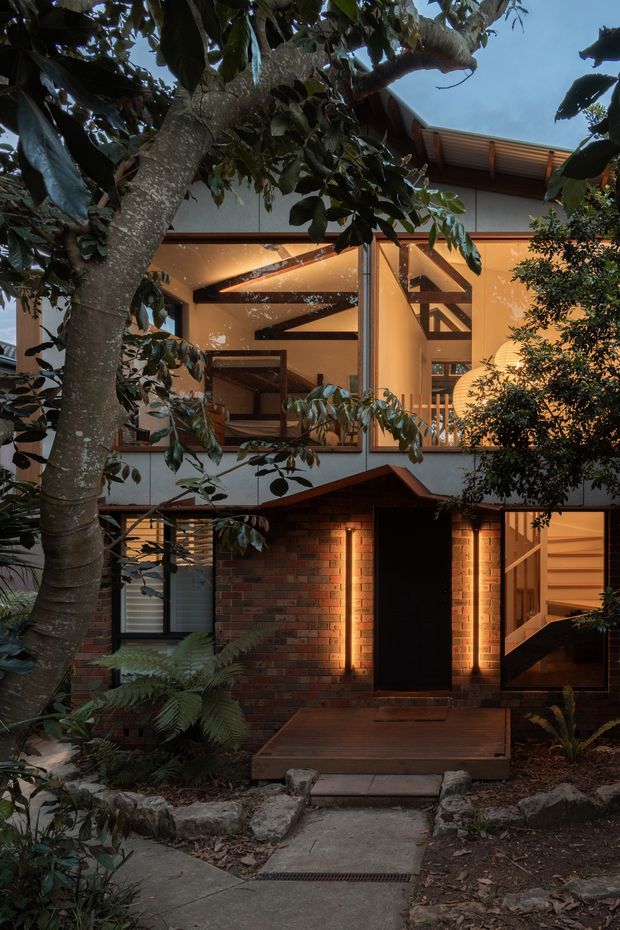 Exterior view of a multi-level residential house set within dense leafy landscaping. The facade combines dark red brick at the lower level with pale rendered walls and extensive timber-framed glazing above. Warm interior lighting reveals exposed timber beams, a stair, and open-plan spaces through the windows. A timber entry deck and concrete pathway lead to the dark front door, with rock edging and ferns integrated into the garden.