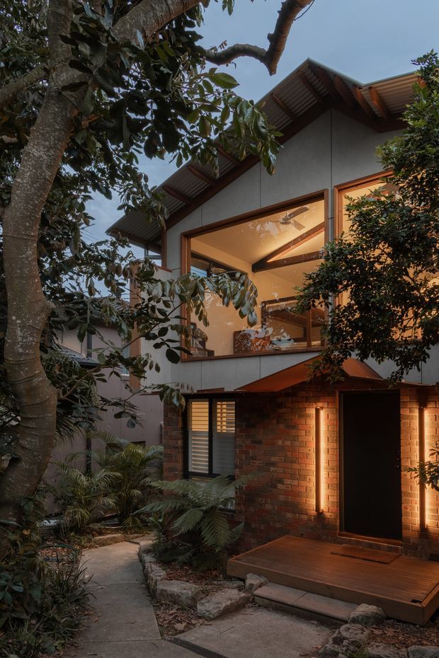 A warmly lit multi-level residential house viewed from a landscaped garden path at dusk. The facade combines red brick at ground level with light grey rendered or panelled walls above, timber-framed glazing, deep roof overhangs, and exposed timber soffits. Large windows reveal a warmly illuminated interior, while a timber deck and dark entry door sit beside dense tropical-style planting and a mature tree.