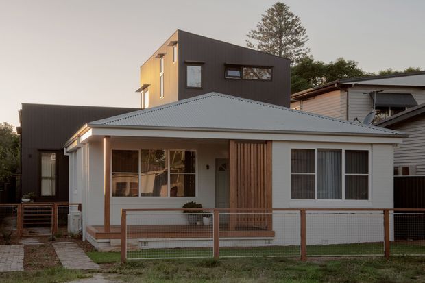 Street-facing view of a compact contemporary two-storey house with a low-pitched corrugated metal roof, dark vertical-clad upper volume, white rendered lower facade, timber-look vertical screen, front porch, large windows, and a simple fenced front yard.