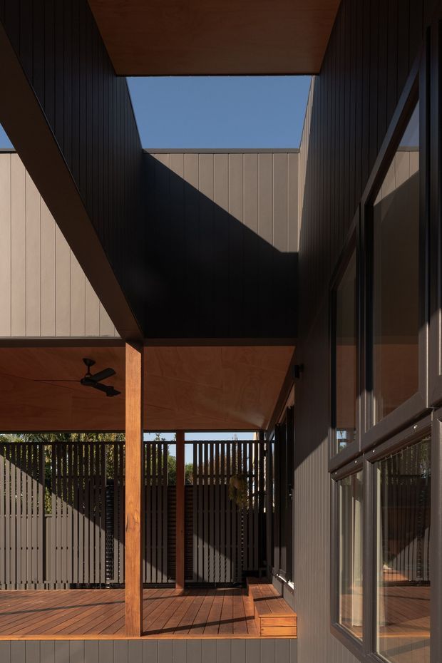 A covered outdoor deck or courtyard-like terrace framed by dark vertical cladding, timber posts, timber soffits, and extensive glazing. Strong angular sunlight creates sharp geometric shadows across the walls and decking, with a ceiling fan and screened privacy fencing visible.