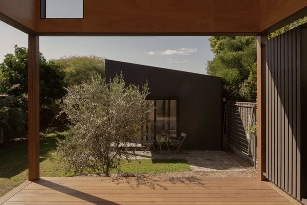 View from a covered timber-framed outdoor deck or terrace toward a compact dark-clad garden pavilion. The scene includes timber decking, warm wood ceiling and posts, vertical dark wall cladding, a glazed pavilion entrance, a small gravel area, and landscaped garden planting.