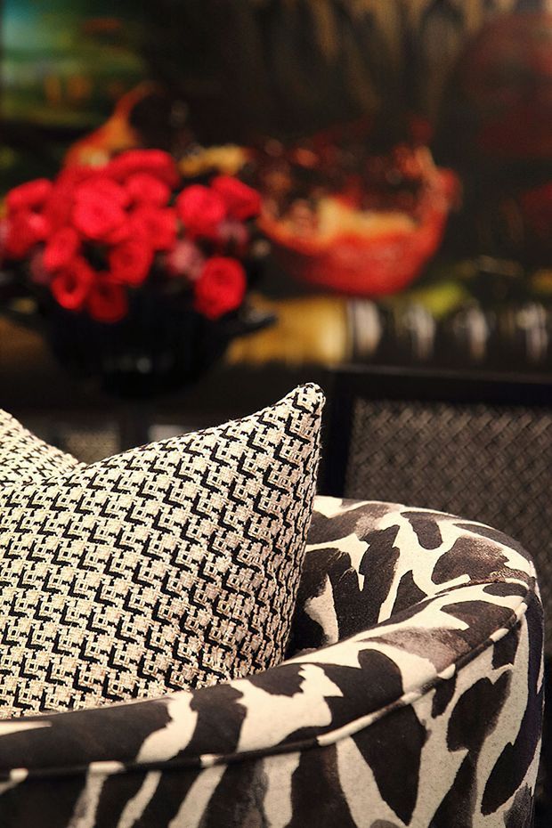 Close-up interior detail of a richly patterned upholstered armchair or lounge chair, featuring black-and-white abstract animal-print fabric, a contrasting cream-and-black geometric cushion, and a dark woven-texture cushion behind it. A blurred arrangement of red flowers and decorative objects sits in the background, creating a warm, dramatic, eclectic atmosphere.