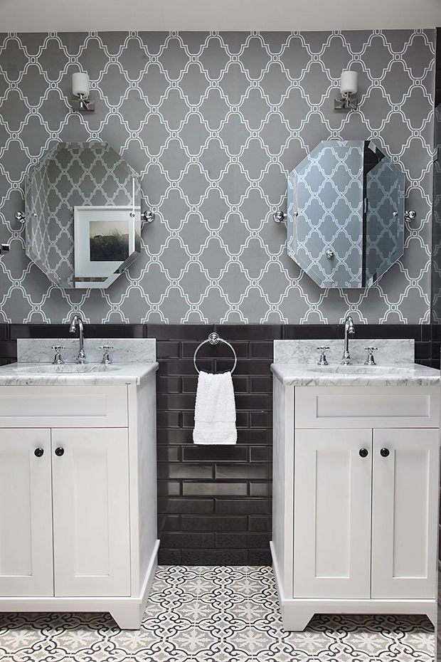 A symmetrical, highly patterned bathroom featuring two white vanity cabinets with marble tops and chrome faucets, a dark glossy subway-tile backsplash, ornate grey-and-white patterned wallcovering, matching patterned floor tiles, and paired wall sconces and mirrors.