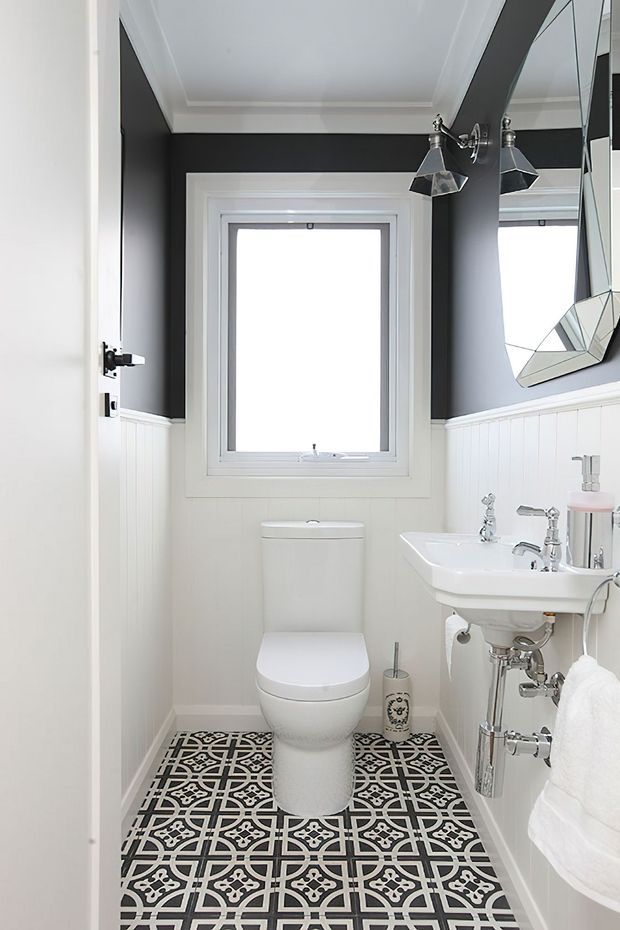 A compact, bright powder room with a white toilet and pedestal-style wall-mounted sink, chrome plumbing and tapware, dark charcoal upper walls, white beadboard wainscoting, white trim, and a bold black-and-white geometric patterned tile floor. A faceted wall mirror and wall-mounted angular light fixtures add decorative detail.