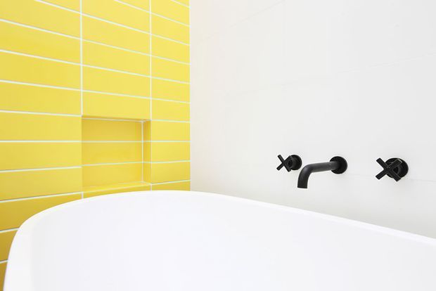 Close-up of a modern bathroom featuring a white freestanding bathtub, bright yellow rectangular tiled wall with a recessed niche, and matte black wall-mounted cross-handle tapware against a white wall.