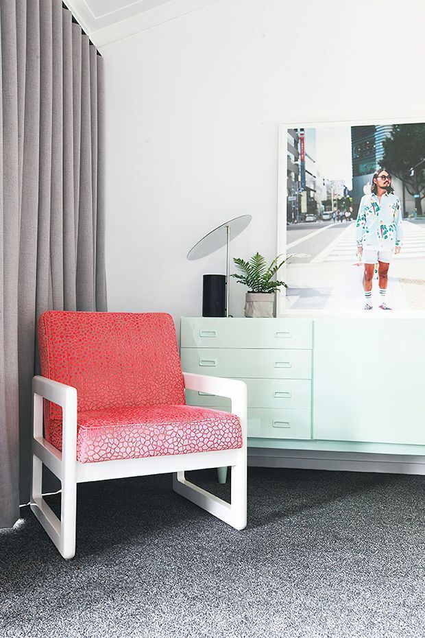A bright, stylish interior corner featuring a white mid-century-inspired lounge chair upholstered in a bold coral-red patterned fabric. It sits beside a white built-in or freestanding drawer cabinet with a black cylindrical lamp, angled oval shade, and small potted fern. Full-height grey curtains, dark grey carpet, white walls and ceiling, and a large framed urban photograph complete the room.