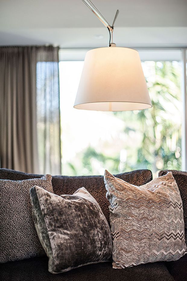 Close-up interior detail of a contemporary living room, featuring a dark upholstered sofa with patterned cushions, a large cream pendant light, full-height windows with sheer curtains, and leafy greenery visible outside. The image emphasizes soft textures, layered neutral tones, and natural light.