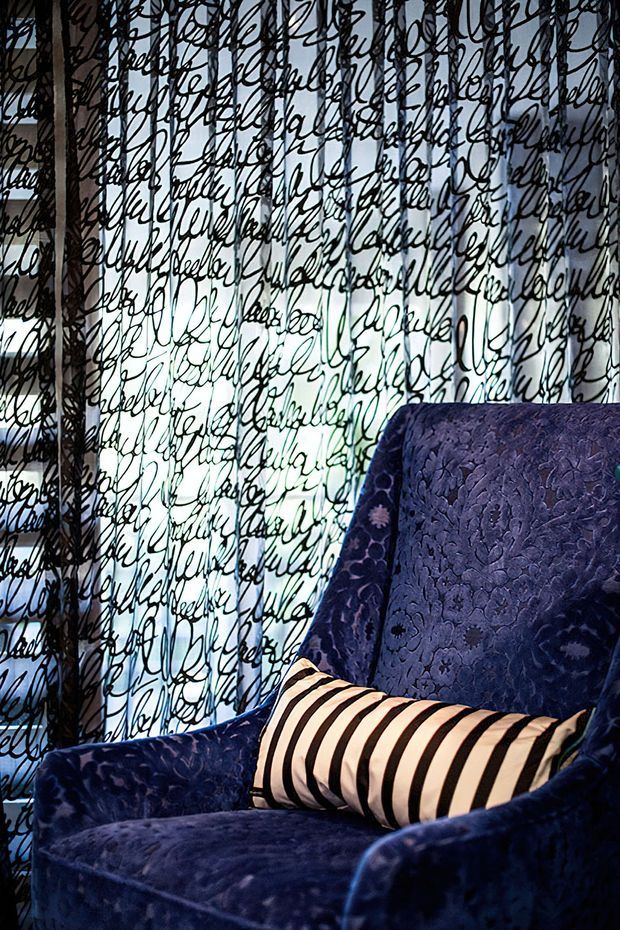 Close-up interior view of a richly upholstered deep navy-blue armchair with floral texture and a cream cushion featuring bold black stripes. Behind it is a translucent or glazed surface covered in dense black handwritten script, creating a dramatic layered backdrop. The scene is dark, intimate, and highly patterned, with strong contrast between the blue upholstery, cream-and-black cushion, and bright background.