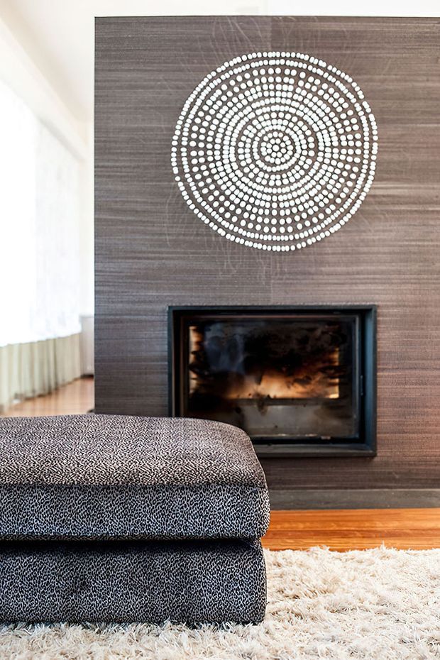 Interior detail of a living room centred on a dark timber-toned feature wall with a built-in open fireplace. A large circular dotted artwork or decorative feature hangs above the fireplace, while a patterned upholstered sofa, timber flooring and a shaggy cream rug occupy the foreground.