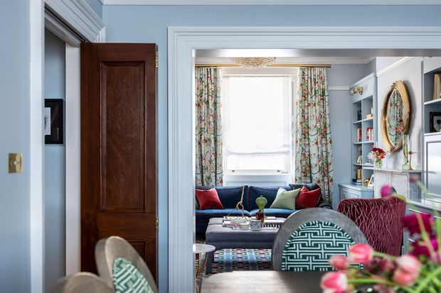 View through a wide, heavily moulded doorway into a richly decorated living room. The room features pale blue walls and trim, a dark blue sofa with colourful cushions, patterned floral curtains, built-in shelving, a large round decorative mirror, and layered patterned textiles. A dark timber panelled door and glimpses of adjacent seating appear in the foreground.