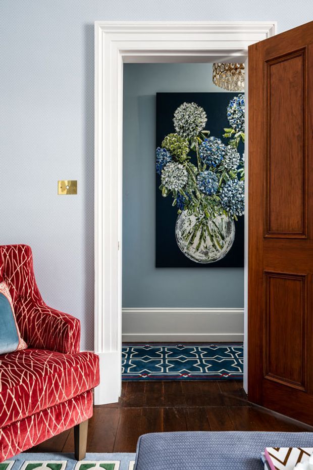 A refined interior view looking through a white-trimmed doorway into a blue-grey hallway. The space features dark timber flooring, a patterned geometric runner, a large floral artwork, a crystal-like pendant light, and a partially visible rich walnut-toned paneled door. In the foreground is a vivid red patterned upholstered armchair and part of a blue-grey upholstered ottoman. The overall composition is colourful, layered, and traditionally detailed.