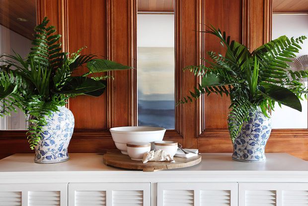 A styled interior vignette featuring two large blue-and-white botanical ceramic planters filled with lush green foliage, positioned on a white built-in sideboard. The backdrop is rich dark timber wall panelling with inset mirrors or reflective panels, while stacked white ceramic bowls and sculptural decorative objects sit centrally on a wooden tray. The composition has a refined, traditional character with strong natural materials and symmetrical styling.