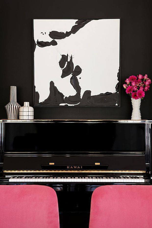 A stylish piano vignette against a black wall, featuring a glossy black upright piano, two bright pink upholstered chairs in the foreground, monochrome abstract artwork, and decorative black-and-white vases with pink flowers. The composition has a dramatic, high-contrast, art-focused interior aesthetic.