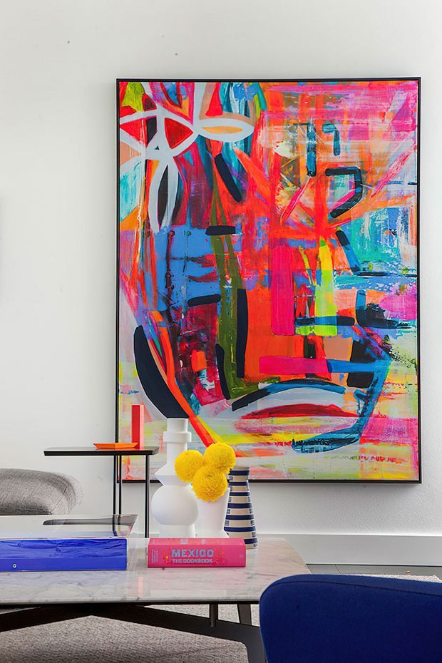 A bright contemporary living room vignette featuring a large, vividly coloured abstract painting in a slim black frame mounted on a white wall. A marble-topped coffee table displays sculptural vases, a striped vessel, a yellow floral arrangement, books and a blue decorative box, with a grey sofa and deep blue seating visible in the foreground.