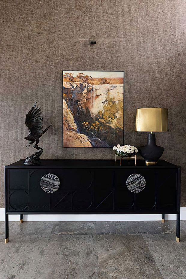A refined interior vignette featuring a black sculptural sideboard against a textured taupe wall. The sideboard has geometric relief detailing, circular stone-like door pulls, and slender brass-toned legs. Above it hangs an abstract landscape artwork beneath a slim linear wall light. A dark sculptural vase with a warm brass shade, a small floral arrangement, and an eagle sculpture complete the arrangement. Large-format grey stone flooring grounds the scene.
