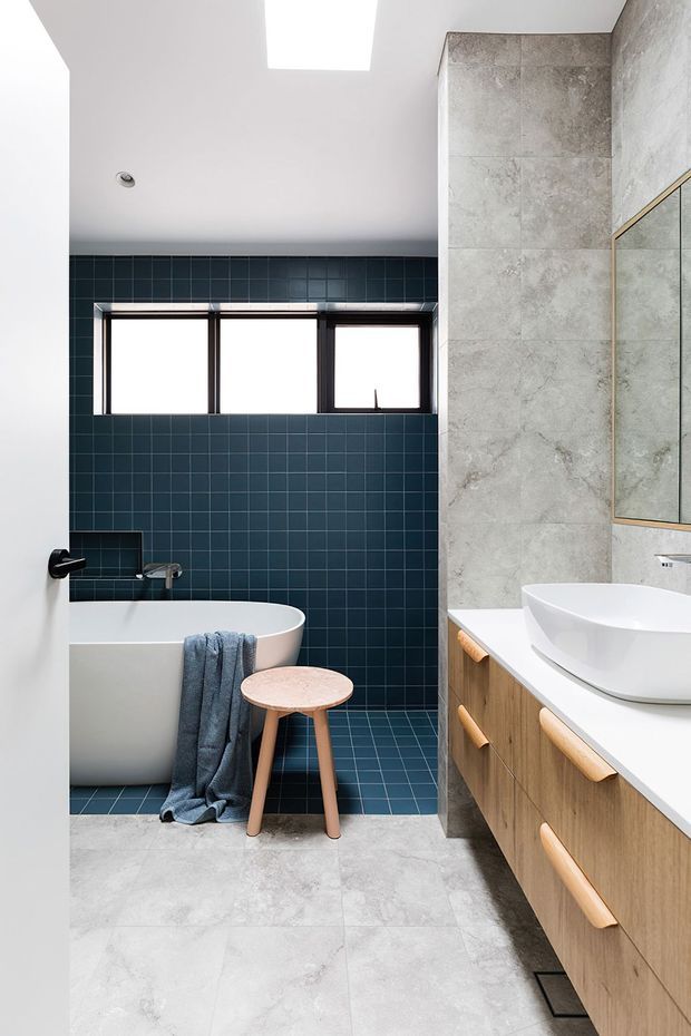 Bright contemporary bathroom with a freestanding white bathtub, wall-mounted vessel sink, timber vanity cabinetry, large grey stone-look wall and floor tiles, and a dark blue tiled feature wall. A high horizontal window and skylight provide natural light, while a frameless glass shower enclosure is visible at right.