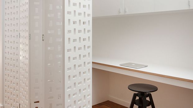 Bright, compact contemporary kitchen or kitchenette with white upper cabinetry, a long white benchtop, integrated cooktop, timber trim, and a dark timber stool. Decorative white perforated partition screens partially divide the space and add geometric detail. The room has warm timber flooring and a clean, minimalist appearance.