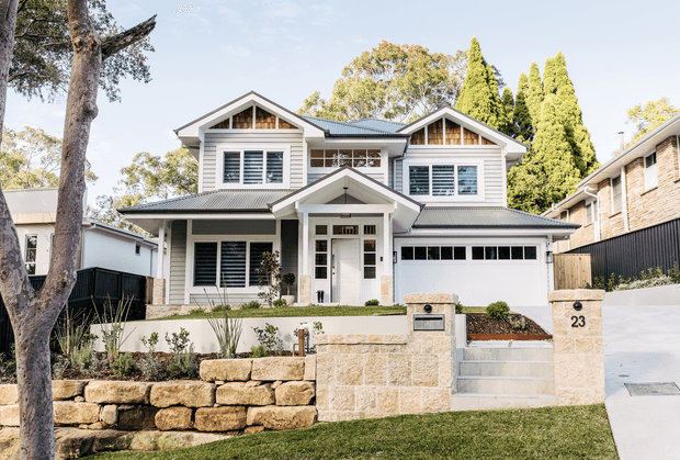 Front exterior view of a large two-storey suburban home with a symmetrical gabled facade, white horizontal weatherboard cladding, dark grey pitched metal roofing, white-framed windows with shutters, a covered entry porch, and an integrated double garage. The property includes a rendered and sandstone retaining wall, landscaped garden beds, front steps, and a concrete driveway.