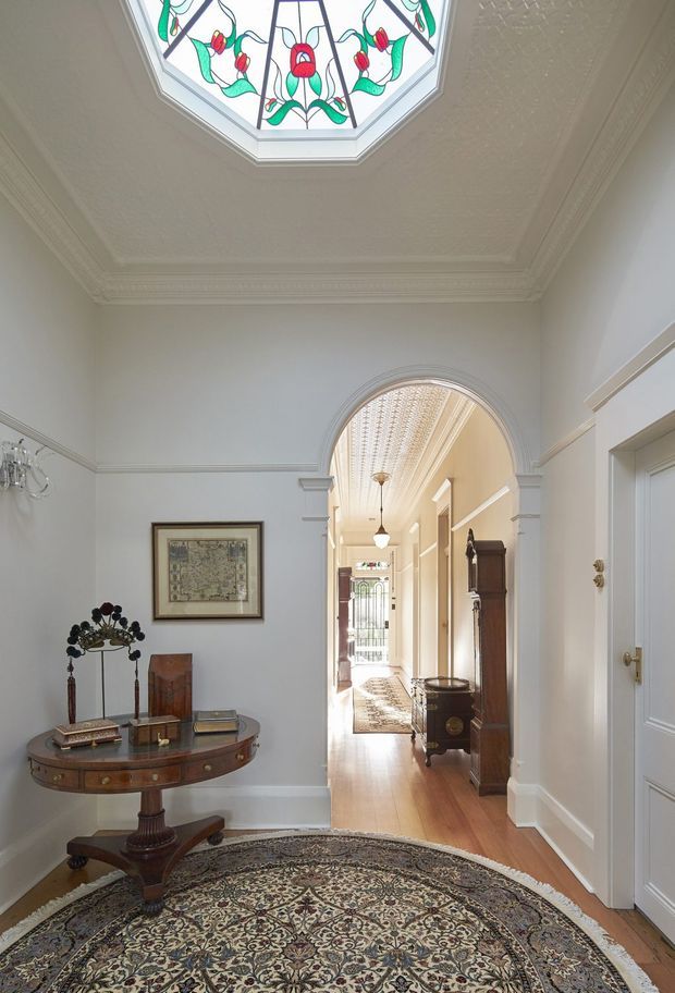 A refined traditional entry hall with pale grey walls, detailed decorative cornices and wall mouldings, an ornate stained-glass ceiling skylight, and a large arched opening leading into a long corridor. The space features polished timber flooring, a round antique timber console table, framed artwork, a tall grandfather clock, period cabinetry, pendant lighting, and a large patterned traditional rug.