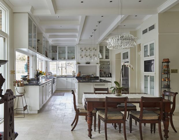 Bright, spacious open-plan kitchen and dining area with white shaker-style cabinetry, glass-front upper cupboards, dark stone benchtops, integrated appliances, and a large central island. A traditional timber dining table and chairs sit beneath an ornate crystal chandelier. The room features tall windows, decorative ceiling beams and mouldings, pale stone-look flooring, and classic architectural detailing.