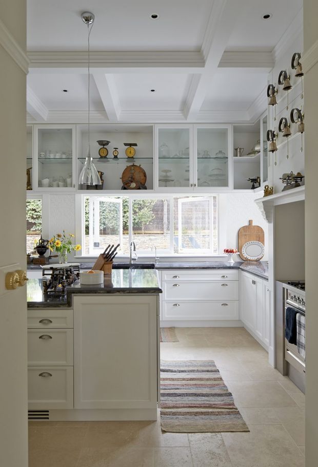 Bright traditional kitchen viewed from the doorway, featuring white shaker-style cabinetry, glass-front upper cabinets, dark stone benchtops, a central island, tiled flooring, and a coffered ceiling with decorative moulding. Large windows provide natural light, while open shelving and display cabinetry add character.