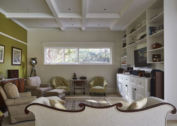 A refined traditional living room with a coffered ceiling, decorative cornices, olive-green and cream walls, upholstered seating, a carved-back sofa in the foreground, and extensive white built-in shelving and cabinetry along one wall. A wide horizontal window provides views to leafy trees, while framed artwork, display objects, and a patterned rug complete the room.