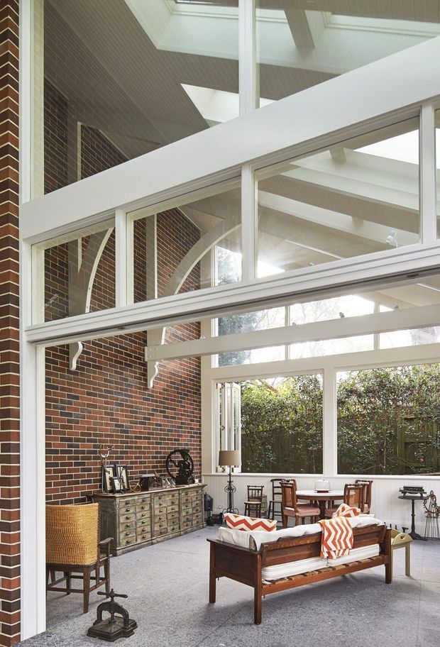 A bright, dramatic double-height living room or sunroom with extensive white-framed glazing and multiple roof skylights. Exposed dark red brick walls contrast with white painted timber beams, ceiling lining, and arch details. The room contains a vintage-style wooden sofa, dining table and chairs, a large drawer-front sideboard, and eclectic furnishings, with greenery visible outside through the windows.