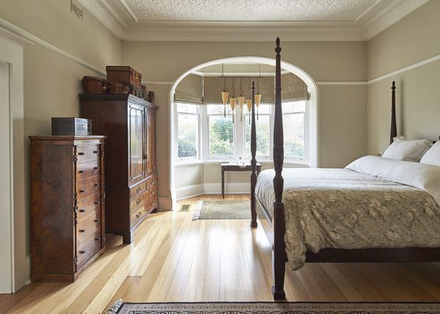 A refined traditional bedroom with a four-poster bed, antique timber chest of drawers and armoire, pale timber flooring, taupe walls with dado-style trim, ornate ceiling detailing, and a large curved bay window. The room is bright and spacious with classic period character and restrained neutral furnishings.