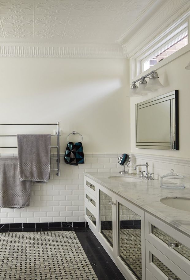A bright, refined traditional bathroom with a long white vanity, marble countertop, undermount basins, mirrored cabinetry, polished chrome fittings, and a large framed mirror. White subway wall tiles are paired with a decorative black-and-white mosaic floor and black border tiles. The room features ornate pressed-tin ceiling panels, detailed cornices, clerestory-style windows, and a heated towel rail.