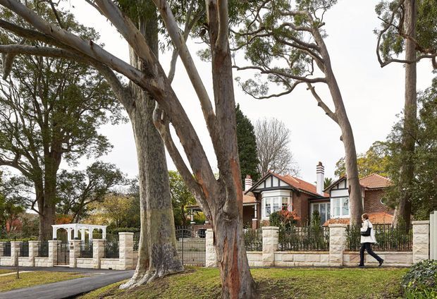 Street-facing view of a traditional suburban home partially screened by mature eucalyptus trees. The house features red terracotta tiled roofs, white rendered walls, black timber-style gable detailing, bay windows, chimneys, and a low pale stone boundary wall with black metal fencing and a gated entrance.