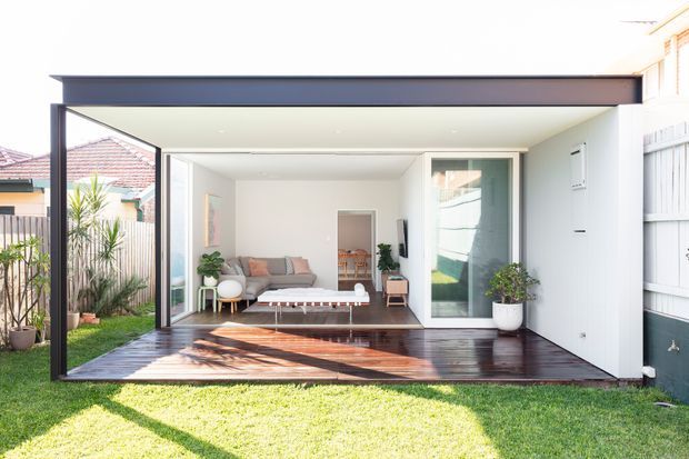 Bright exterior view of a contemporary single-storey rear extension opening onto a timber deck and lawn. The pavilion-like addition has a broad dark-framed opening, white walls, large glazing, a flat roof, and a compact living area visible inside with a sofa, coffee table, plants, and built-in storage. Neighbouring residential structures are visible around the edges.